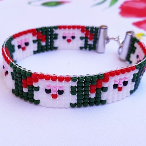 Op de afbeelding: Een groene, witte en rode kralen armband met een patroon van cartoon gezichten. De armband heeft een zilveren sluiting en ketting.