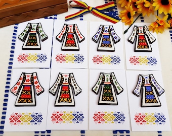 Pode incluir: Oito pinos de madeira com desenhos bordados coloridos de roupas tradicionais romenas. Cada pino é exibido em um cartão branco com uma borda estampada em vermelho, amarelo e azul.
