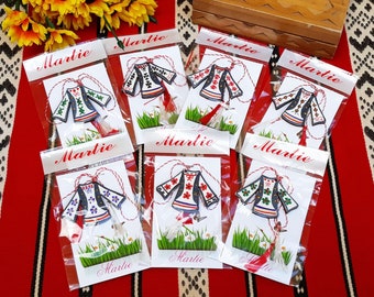 Pode incluir: Oito pequenos amuletos martie romenos feitos à mão, coloridos, cada um com um design tradicional de blusa bordada. Os amuletos são embalados individualmente em cartões brancos com a palavra "Martie" impressa neles.