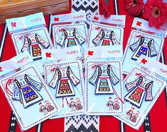 Pode incluir: Um conjunto de oito trajes folclóricos romenos coloridos tradicionais, cada um com bordados intrincados e um cordão vermelho anexado. Os trajes são exibidos em cartões brancos com a palavra "martie" impressa neles.