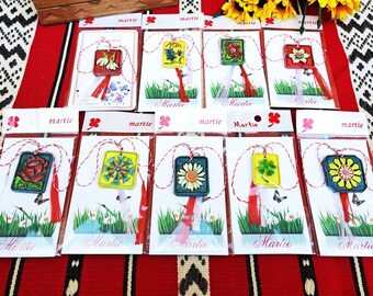 Pode incluir: Nove amuletos martisor coloridos, cada um com um design floral diferente, são exibidos em cartões individuais com a palavra "martie" impressa neles. Os amuletos estão presos a cordas vermelhas e brancas.