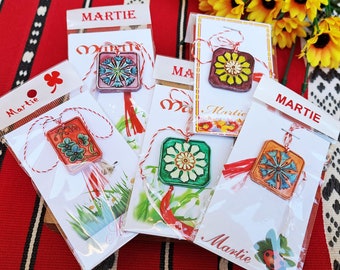 Pode incluir: Cinco pequenos amuletos quadrados coloridos com desenhos florais, cada um em um cartão branco com a palavra "Martie" impressa nele. Os amuletos estão presos a cordas vermelhas e brancas.