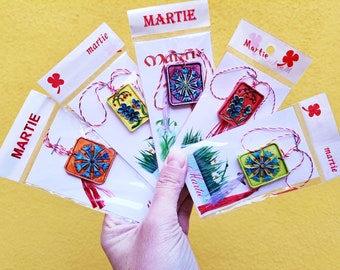 Pode incluir: Seis amuletos quadrados coloridos com desenhos florais e um cordão vermelho atado. Os amuletos estão embalados em sacos de plástico transparentes com a palavra "Martie" impressa neles.