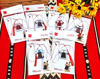 Pode incluir: Seis amuletos martisor coloridos com cordão vermelho e branco. Cada amuleto apresenta um traje tradicional romeno com bordados. Os amuletos são embalados em cartões brancos com a palavra "Martie" impressa neles.