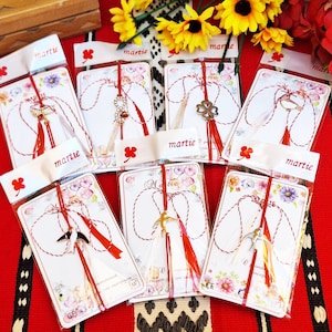 Op de afbeelding: Een verzameling Martisor-bedels, elk verpakt op een witte kaart met bloemaccenten en een rood-wit touwtje. De bedels hebben verschillende vormen, waaronder een sleutel, een klavertjevier en een ster, met rode kwastjes.