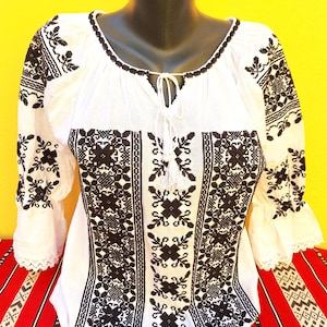 Könnte beinhalten: Weiße Bluse mit schwarzer Blumenstickerei. Das traditionelle Kleidungsstück hat einen runden Ausschnitt, lange Ärmel mit Spitzensaum und einen Kordelverschluss. Die Bluse wird auf einer Schaufensterpuppe präsentiert.