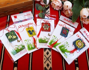 Pode incluir: Cinco amuletos quadrados coloridos com desenhos florais e borlas vermelhas. Cada amuleto é embalado em um saco de plástico transparente com um cartão branco que diz "Martie" em uma fonte decorativa.