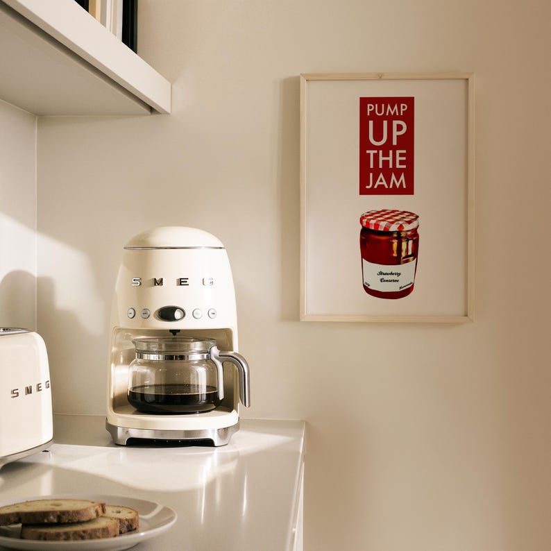 Puede incluir: Cafetera retro de color crema con una jarra de vidrio llena de caf&eacute;. Un estampado enmarcado con las palabras "PUMP UP THE JAM" y una ilustraci&oacute;n de un tarro de mermelada de fresa cuelga de la pared. Tambi&eacute;n se ven una tostadora y un plato de tostadas.