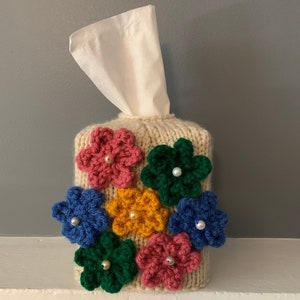 Könnte beinhalten: Ein gestrickter Taschentuchbox-Bezug mit beigefarbenem Hintergrund und bunten gehäkelten Blumen. Die Blumen sind rosa, blau, grün und gelb mit weißen Perlen in der Mitte.