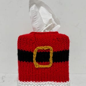 Puede incluir: Una funda rectangular para caja de pañuelos hecha de hilo rojo, negro y blanco. La funda presenta un cuerpo de punto rojo con una banda negra horizontal en el centro, un cuadrado de hilo amarillo que representa una hebilla y un borde blanco en la base.