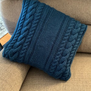 Può includere: Un cuscino blu lavorato a maglia con un motivo a treccia. Il cuscino è seduto su un divano beige.