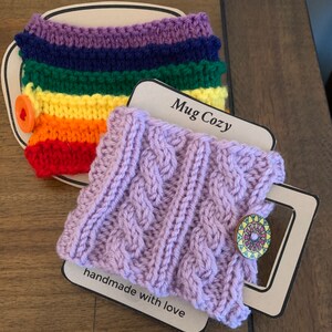 Op de afbeelding: Twee gebreide bekerwarmers. E&eacute;n is een regenboog gestreept ontwerp met een rode knoop. De andere is een paarse kabel gebreid ontwerp met een kleurrijke knoop. Beide bekerwarmers liggen op een bruin oppervlak met een label waarop "Mug Cozy" en "handgemaakt met liefde" staat.