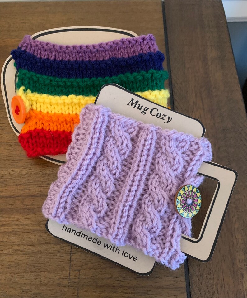 Op de afbeelding: Twee gebreide bekerwarmers. E&eacute;n is een regenboog gestreept ontwerp met een rode knoop. De andere is een paarse kabel gebreid ontwerp met een kleurrijke knoop. Beide bekerwarmers liggen op een bruin oppervlak met een label waarop "Mug Cozy" en "handgemaakt met liefde" staat.