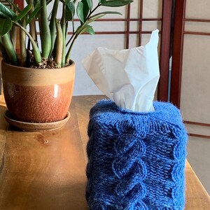 Könnte beinhalten: Ein blauer gestrickter Taschentuchbox-Bezug mit einem Zopfmuster. Der Bezug liegt auf einem Holztisch mit einer Pflanze in einem Terrakotta-Topf im Hintergrund.