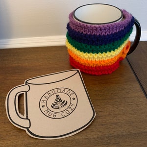 Op de afbeelding: Een regenboogkleurige gehaakte mokwarmer met een knoopsluiting. De mokwarmer zit op een witte keramische mok. Op de voorgrond ligt een bruine papieren uitsnede van een mok met de tekst "Handmade Mug Cozy".