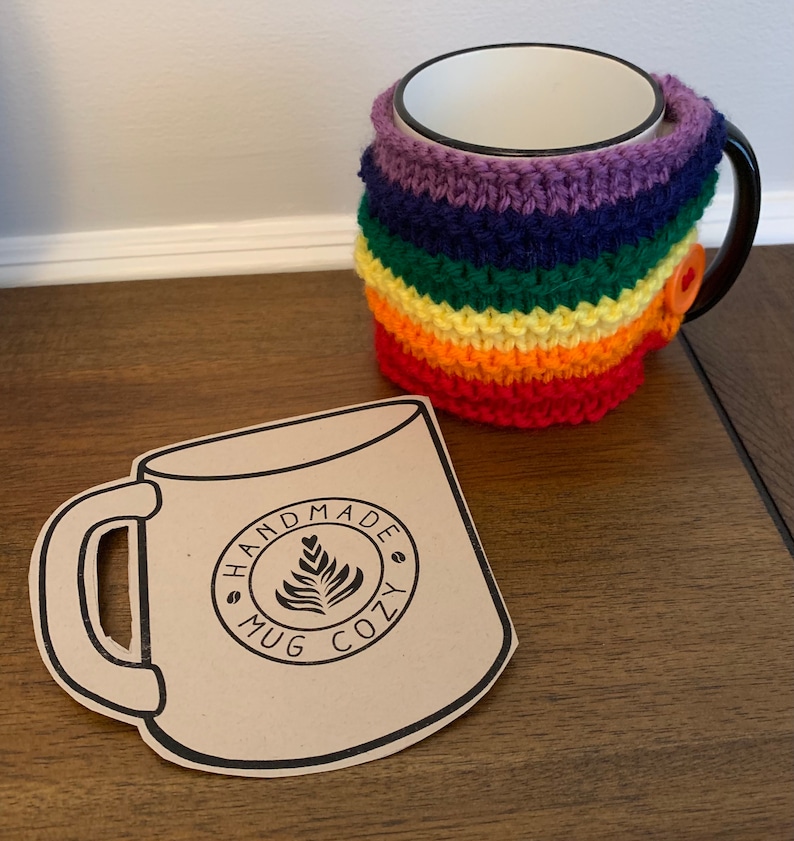 Op de afbeelding: Een regenboogkleurige gehaakte mokwarmer met een knoopsluiting. De mokwarmer zit op een witte keramische mok. Op de voorgrond ligt een bruine papieren uitsnede van een mok met de tekst "Handmade Mug Cozy".