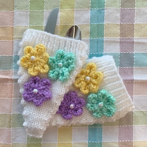 May include: White knitted utensil holders with crocheted flowers in yellow, purple, and green. Each flower has a pearl bead in the center.