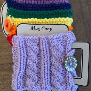 Op de afbeelding: Een paarse gebreide mokwarmer met een kabelbreipatroon en een houten knoop met een kleurrijk ontwerp. De mokwarmer is handgemaakt met liefde en heeft de tekst "Mug Cozy" op het label.
