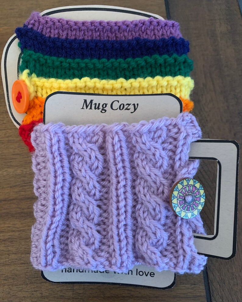 Op de afbeelding: Een paarse gebreide mokwarmer met een kabelbreipatroon en een houten knoop met een kleurrijk ontwerp. De mokwarmer is handgemaakt met liefde en heeft de tekst "Mug Cozy" op het label.