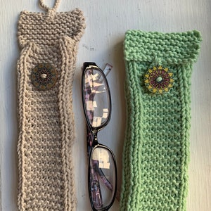 Puede incluir: Dos estuches para gafas de punto, uno beige y otro verde, con botones decorativos. El estuche beige tiene un lazo para colgar.