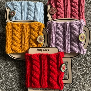 Op de afbeelding: Vier gebreide bekerwarmers in verschillende kleuren: lichtblauw, geel, paars en rood. Elke bekerwarmer heeft een gevlochten patroon en een houten knoop met een bloemenmotief. De bekerwarmers worden weergegeven op een bruine achtergrond met de tekst "Mug Cozy" en "handmade with love".