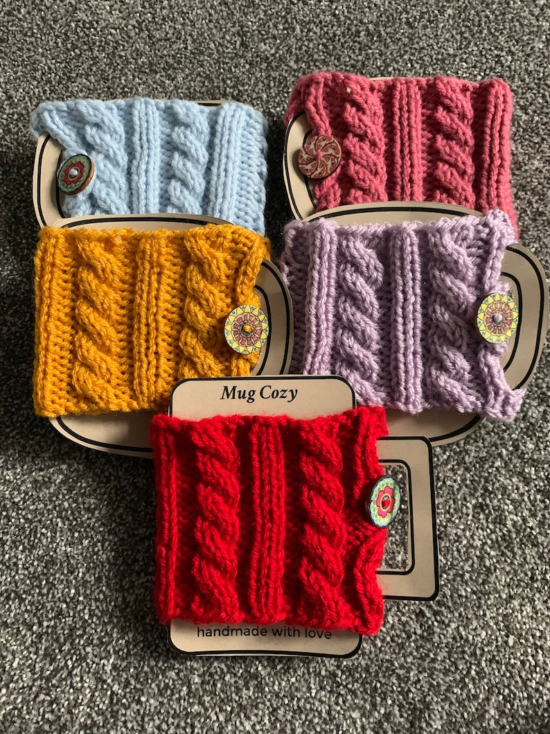 Op de afbeelding: Vier gebreide bekerwarmers in verschillende kleuren: lichtblauw, geel, paars en rood. Elke bekerwarmer heeft een gevlochten patroon en een houten knoop met een bloemenmotief. De bekerwarmers worden weergegeven op een bruine achtergrond met de tekst "Mug Cozy" en "handmade with love".