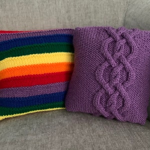 Può includere: Due cuscini di maglia, uno con un motivo a righe arcobaleno e l'altro con un motivo a treccia viola.