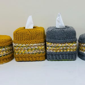 May include: Four knitted tissue box covers and matching small containers. Two are mustard yellow, two are gray. Each has a band of patterned yarn in white, green, and yellow. The tissue boxes have white tissues sticking out.