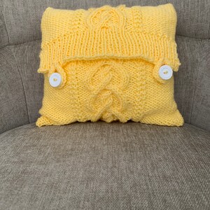 Pu&ograve; includere: Un cuscino giallo lavorato a maglia con un motivo a treccia e due bottoni bianchi. Il cuscino &egrave; seduto su un divano grigio.