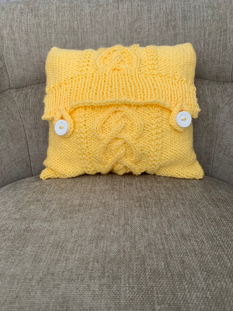 Pu&ograve; includere: Un cuscino giallo lavorato a maglia con un motivo a treccia e due bottoni bianchi. Il cuscino &egrave; seduto su un divano grigio.