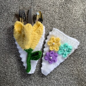 Pode incluir: Um conjunto de dois porta-talheres de malha. Um porta-talheres tem a forma de uma flor amarela com folhas verdes e o outro é branco com três flores de crochê em amarelo, roxo e verde.