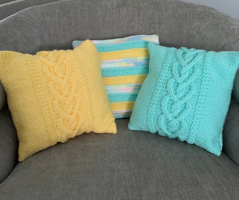 Pu&ograve; includere: Tre cuscini da divano in maglia con motivi a treccia. I cuscini sono gialli, blu chiaro e con un motivo a righe multicolore.
