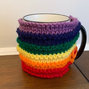 Op de afbeelding: Een regenboogkleurige gebreide mokwarmer met een knoopsluiting. De warmer is gemaakt van zacht, kleurrijk garen en is perfect om je handen warm te houden terwijl je geniet van je favoriete drankje.