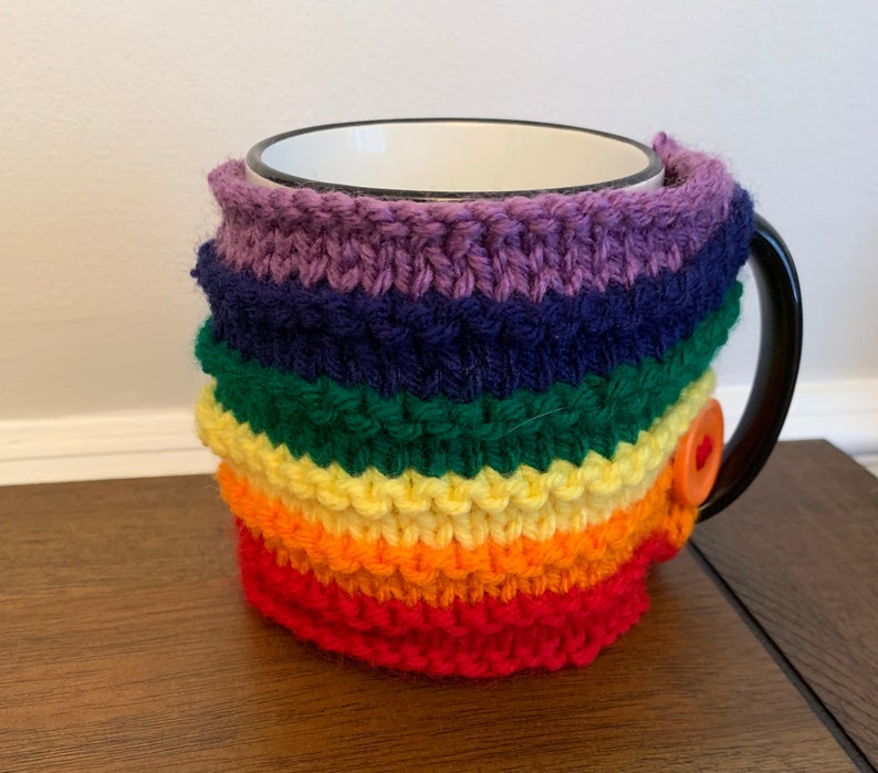 Op de afbeelding: Een regenboogkleurige gebreide mokwarmer met een knoopsluiting. De warmer is gemaakt van zacht, kleurrijk garen en is perfect om je handen warm te houden terwijl je geniet van je favoriete drankje.
