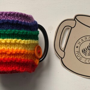 Op de afbeelding: Gebreide mokwarmer met regenboogstrepen en een knoopsluiting. De mokwarmer is handgemaakt en heeft een label met de tekst "Handmade Mug Cozy".