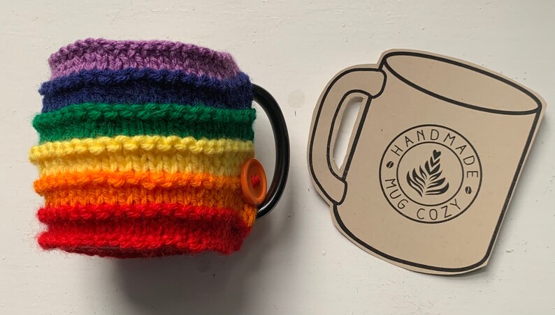 Op de afbeelding: Gebreide mokwarmer met regenboogstrepen en een knoopsluiting. De mokwarmer is handgemaakt en heeft een label met de tekst "Handmade Mug Cozy".