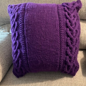 Può includere: Un cuscino di maglia viola con un motivo a treccia sui bordi.