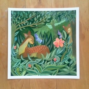 May include: A square art print featuring a painted scene of a brown spotted fawn resting in a lush green forest. The composition includes dense foliage, a large pink flower, blue blossoms, and a second deer partially visible behind a tree trunk.