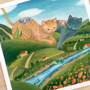 May include: A square art print featuring a painting of a large orange tabby cat and a large grey cat lounging over a green hilly landscape with a blue river, small houses, and orange flowers. The cats have floral patterns on their backs.