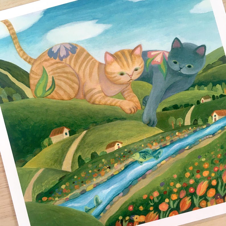 May include: A square art print featuring a painting of a large orange tabby cat and a large grey cat lounging over a green hilly landscape with a blue river, small houses, and orange flowers. The cats have floral patterns on their backs.