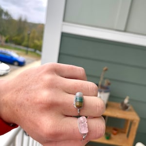Peut inclure: Deux bagues en fil de fer avec des pierres naturelles. La première bague présente une pierre multicolore avec des rayures vertes, brunes et blanches. La deuxième bague présente une pierre rose.