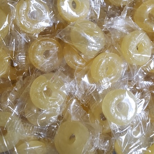 May include: A close-up shot of a pile of yellow, ring-shaped candies individually wrapped in clear plastic. The candies have a hole in the center and appear to be hard candies. The wrappers are slightly crinkled.