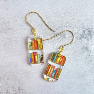 May include: A pair of gold earrings with a clear rectangular charm featuring a miniature bookshelf filled with colorful books.