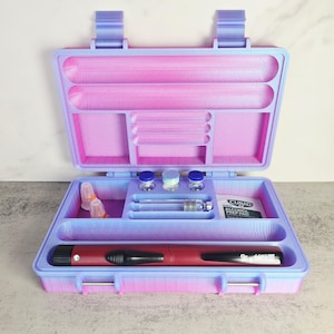 May include: A two-tone pink and blue medical travel case, open to reveal compartments and medical supplies. The case contains vials, syringes, and an alcohol prep pad. A red and black medical pen is also visible.