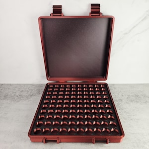 May include: Open, burgundy-colored case with a grid of small, round compartments. The case is rectangular with a hinged lid and two clasps. The interior is a darker shade of burgundy. The case is likely designed for organizing small items.