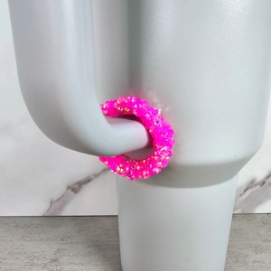 May include: A pink beaded ring with iridescent accents. The ring is wrapped around the handle of a white mug.
