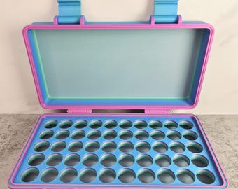 May include: An open, multi-colored case with a blue interior and a pink and blue exterior. The case has a grid of circular holes in the bottom half. The top half is open and has two blue clasps. The case is sitting on a gray surface.