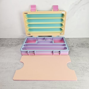 May include: Open, pastel-colored organizer with a hinged lid and multiple compartments. The interior features a gradient of colors, including pink, purple, and blue. The exterior is a light yellow with pink clasps. A matching pink tray sits in front.