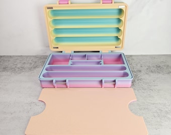 May include: Open, pastel-colored organizer with a hinged lid and multiple compartments. The interior features a gradient of colors, including pink, purple, and blue. The exterior is a light yellow with pink clasps. A matching pink tray sits in front.