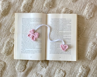 May include: Close-up of an open book with a pink crocheted bookmark shaped like a heart. The book is open to a page with text about a fictional character named "The Peacekeeper".